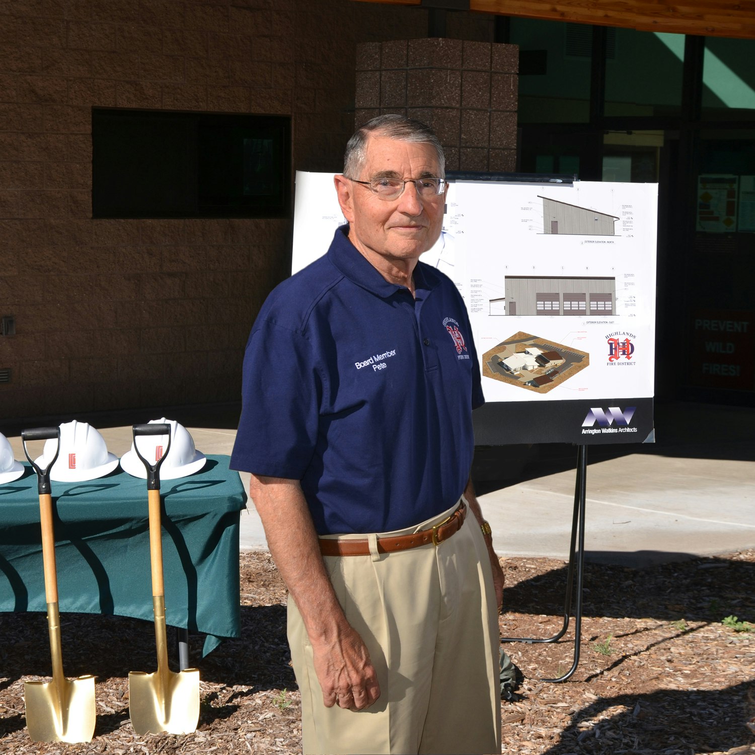 A man in a polo shirt stands near ceremonial shovels and a project display board, possibly at a groundbreaking event.