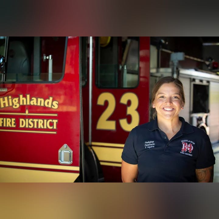 A smiling person in firefighter paramedic uniform in front of a fire truck marked "23".
