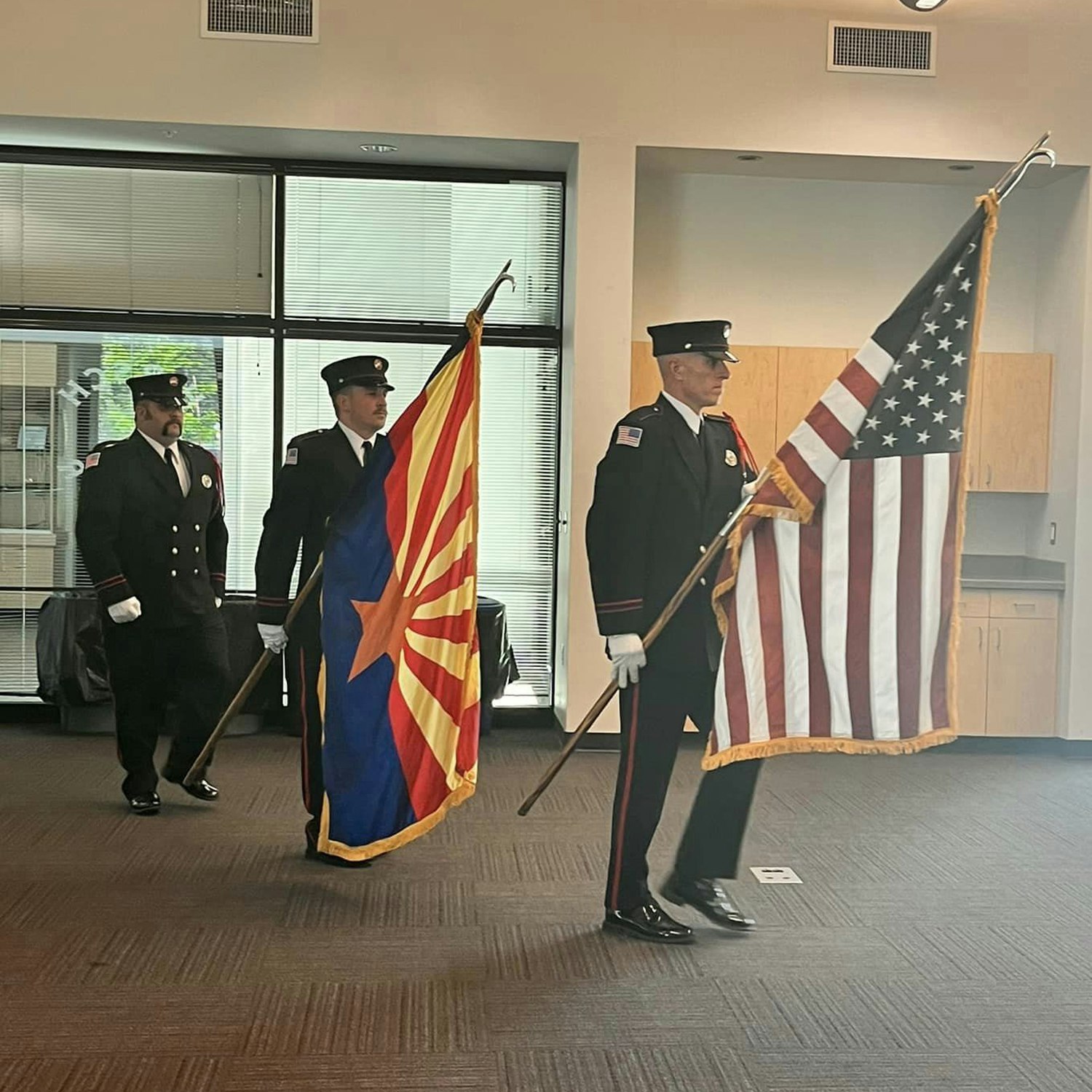 Firefighters in uniform marching with the American flag and another flag indoors.