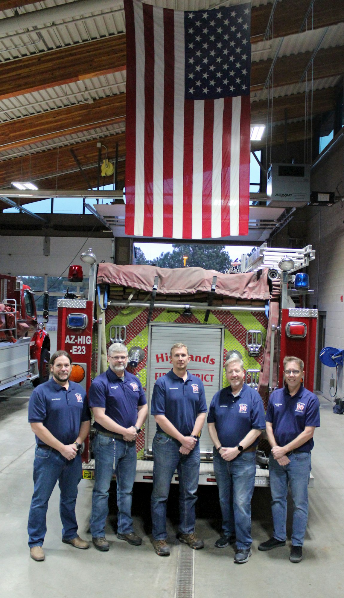 Five firefighters in matching uniforms stand in front of a fire truck inside a fire station. An American flag hangs from the ceiling above them.