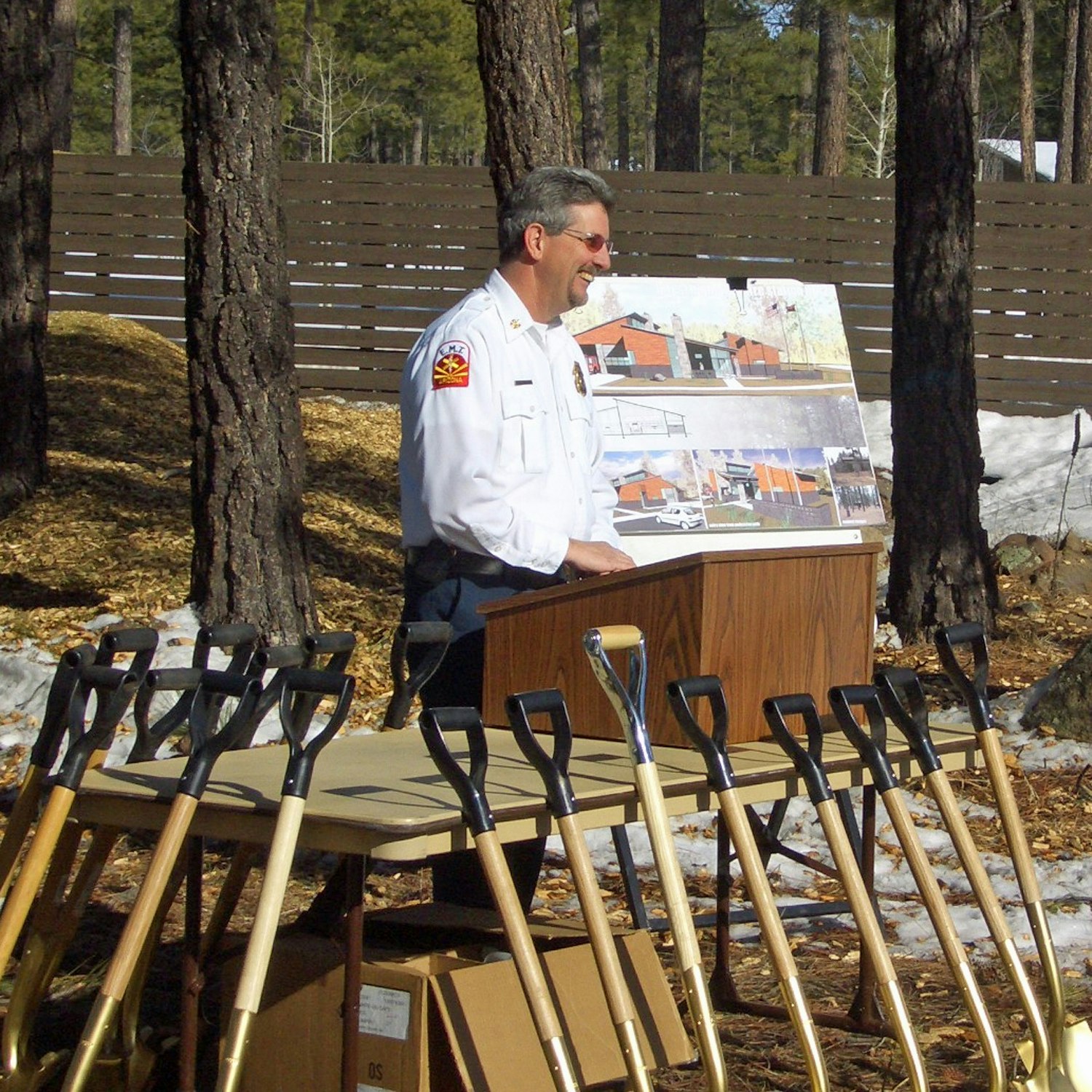 A man in a white uniform presenting at a podium with an image board, multiple ceremonial shovels lined up in front, in an outdoor setting.