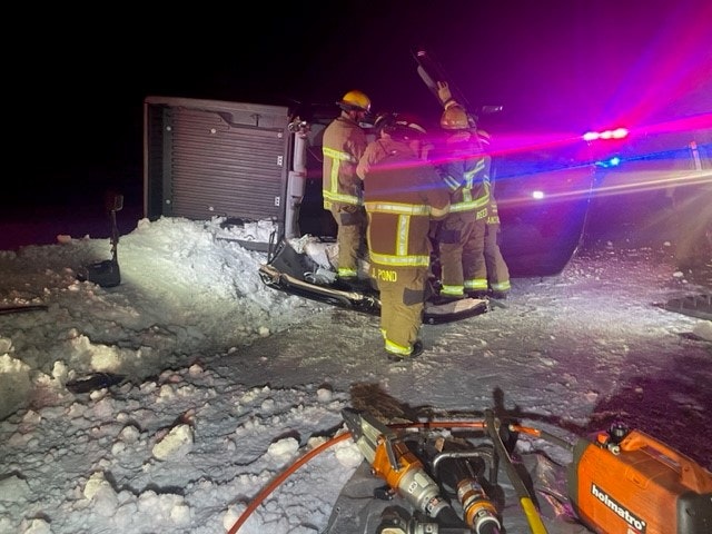 Firefighters near an overturned vehicle at night with emergency lights and rescue equipment on snowy ground.