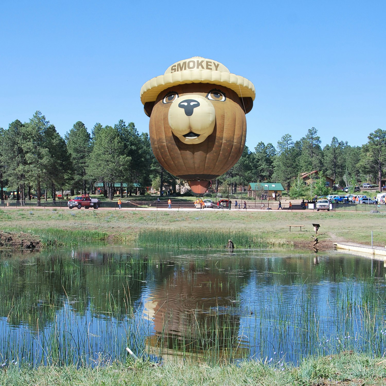 Hot air balloon shaped like Smokey Bear floating near a pond with reflection, surrounded by trees and people.