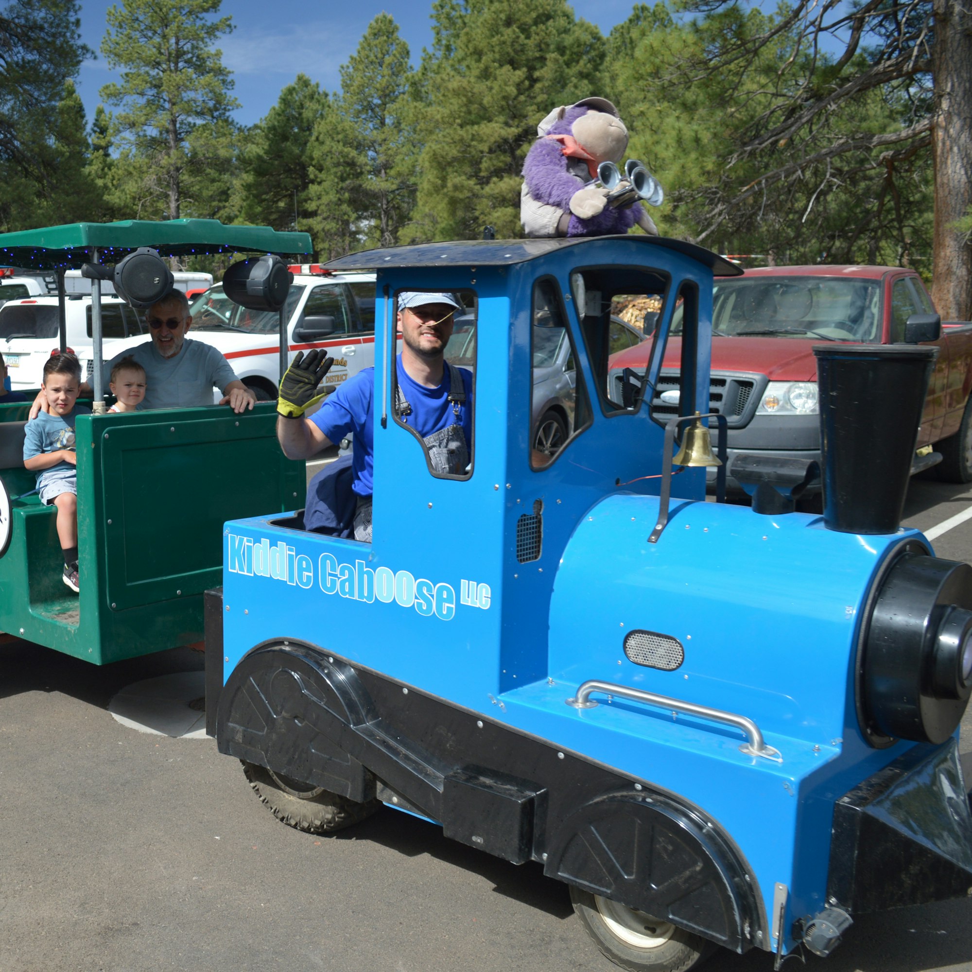 Blue kiddie train ride with passengers, including a plush toy, driven by a man in sunglasses.