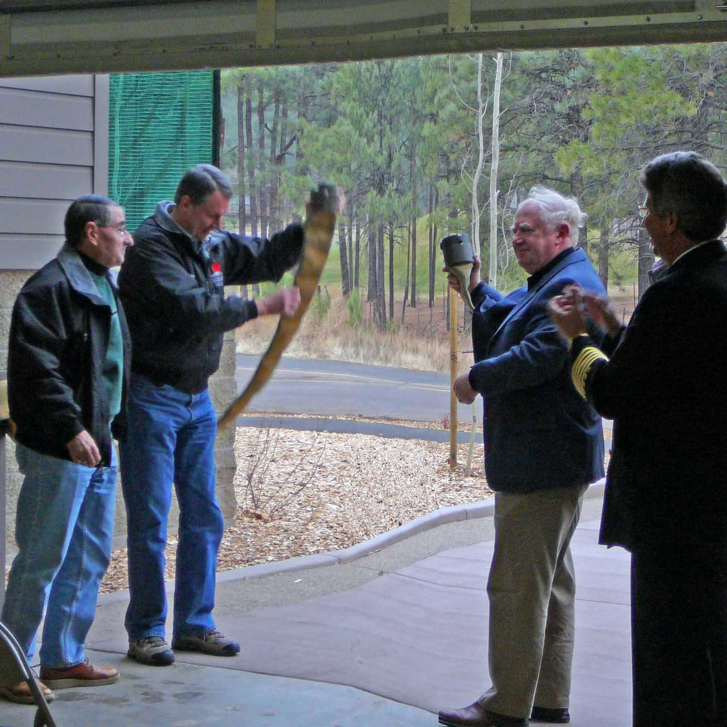 Four men under a porch, one swinging a golf club, others watching. Trees and a driveway in the background.