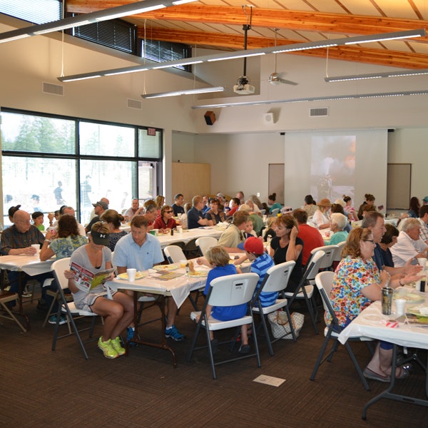 A group of people seated at tables in a well-lit hall, possibly attending an event or a community gathering.