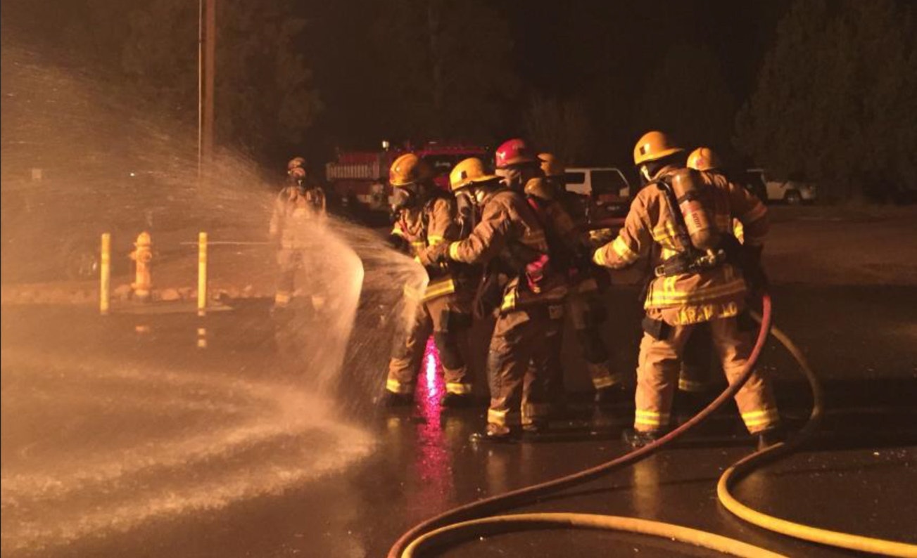 Firefighters operating a hose at night, with water spraying and emergency vehicles visible in the background.