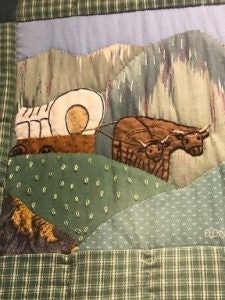 A quilted image of oxen pulling a covered wagon through hilly terrain with a mountain backdrop.