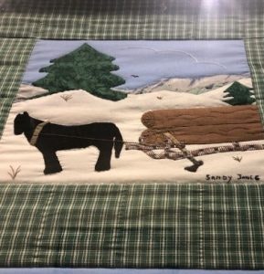 A textile artwork depicting a horse pulling logs in a snowy landscape with trees, signed "Sandy Jones."