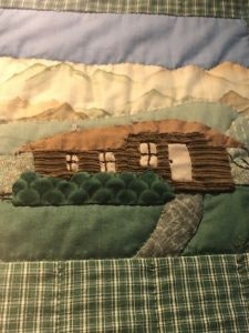 A quilted fabric art piece depicting a log cabin with green bushes, mountains in the background, and a checkered border.