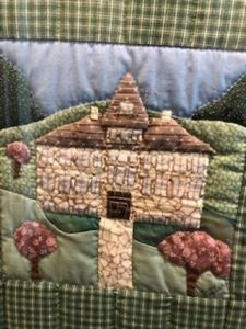 A quilted design featuring a house with a triangular roof, surrounded by trees and hills in various patterned fabrics.