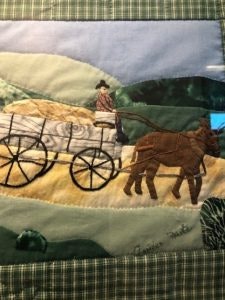 A quilt patch depicting a person in a wagon pulled by two horses, set against a green and blue landscape background.