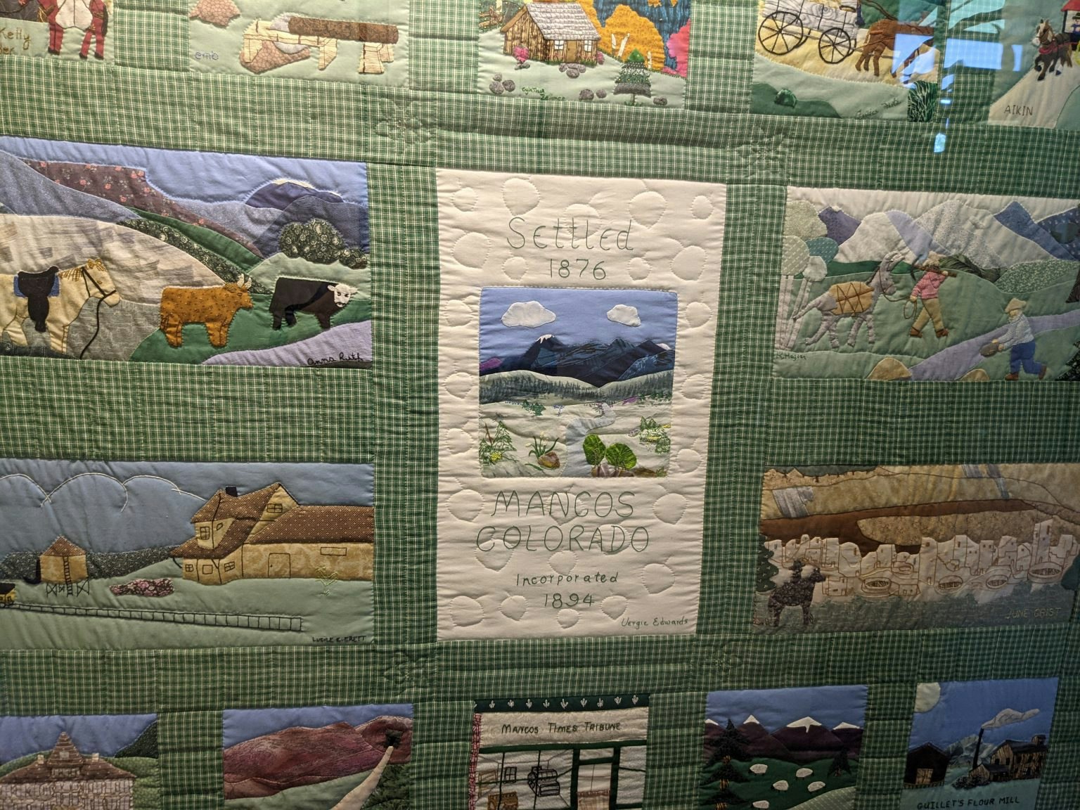 Quilt depicting scenes from Mancos, Colorado, including mountains, animals, and historical elements. Center text: "Settled 1876, Mancos, Colorado."