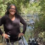 A person smiling outdoors by a river, with trees in the background, holding a dog on a leash.