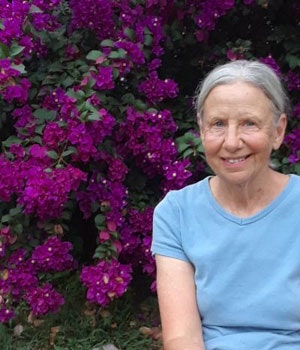 A person in a blue shirt is sitting near vibrant purple flowers, smiling at the camera.