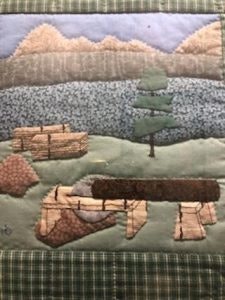 A quilt with a woodland scene featuring mountains, logs, a tree, and a riverbank against a plaid border.