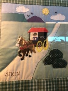 Fabric art depicting a horse-drawn carriage labeled "Doc" on a road with a sun, clouds, mountains, and the name "Aikin."