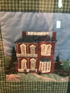 Quilted artwork featuring a red and white house with trees, set against a blue sky with a green plaid border.