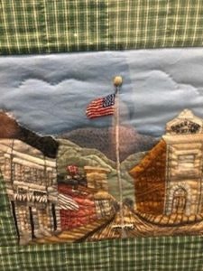 Quilted scene of a town with buildings, mountains, and an American flag on a pole, set against a plaid background.