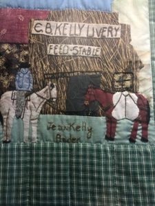 A patchwork quilt depicting a livery feed stable with horses. Text reads "E.B. Kelly Livery Feed Stable" and "Jean Kelly Baker."