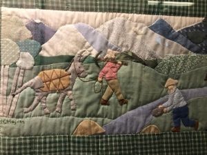 A fabric quilt depicts two people with a pack animal walking through a mountainous landscape.