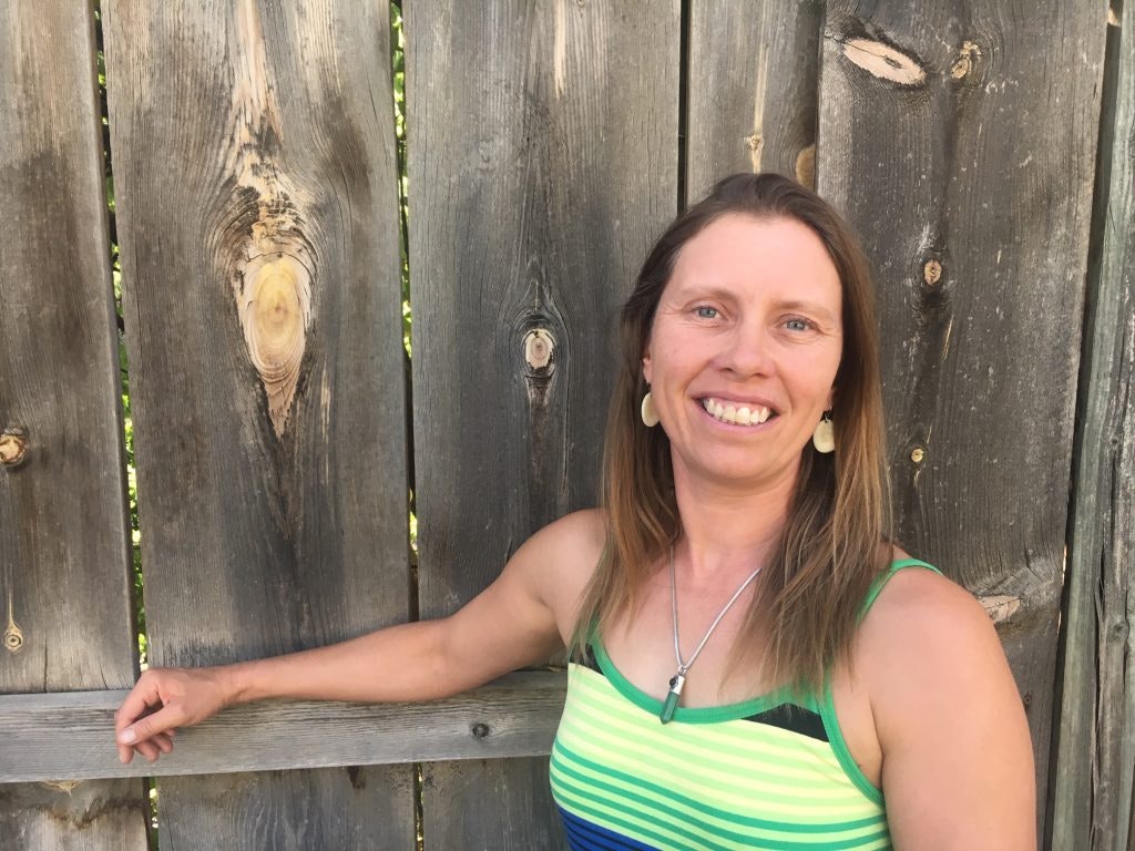 A smiling woman stands outdoors against a wooden fence wearing a colorful striped tank top.