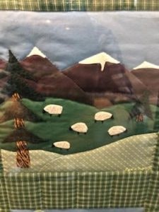 A quilt featuring a mountain landscape with trees and sheep on green hills.