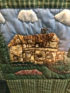 A quilted fabric panel depicting a rustic house with a thatched roof, trees, and clouds.