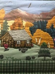A quilted scene with a cabin, trees, mountains, and a bird in the sky.