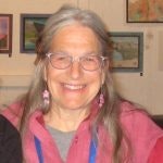 An individual with long hair, glasses, and a pink jacket smiles in a room with framed artworks on the walls.