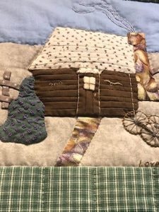 A patchwork quilt depicting a cabin with a textured roof, chimney, tree, and multicolored path, set against a blue and green background.