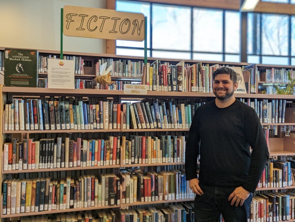 Man standing by a library's fiction section, with bookshelves full of colorful books and a "Fiction" sign on top.