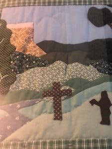 Patchwork quilt with landscape design: hills, trees, and two crosses on a grid-patterned border.