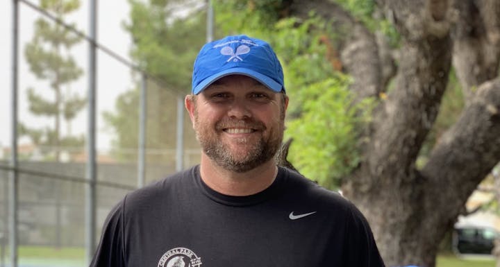 The image features a smiling man with a beard, wearing a blue cap and black shirt, standing near a tennis court and trees.