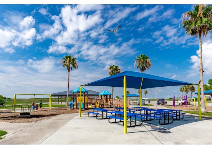 Outdoor playground with picnic tables, swings, slides, blue canopies, palm trees, and a clear sky.