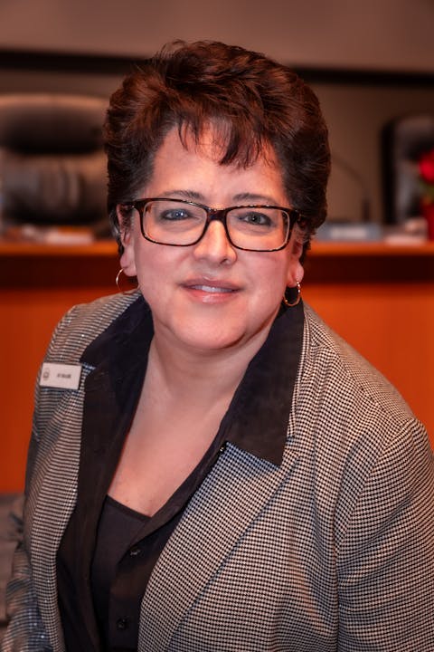A smiling woman wearing glasses and a patterned blazer, posed against a blurred background, possibly in a professional setting.