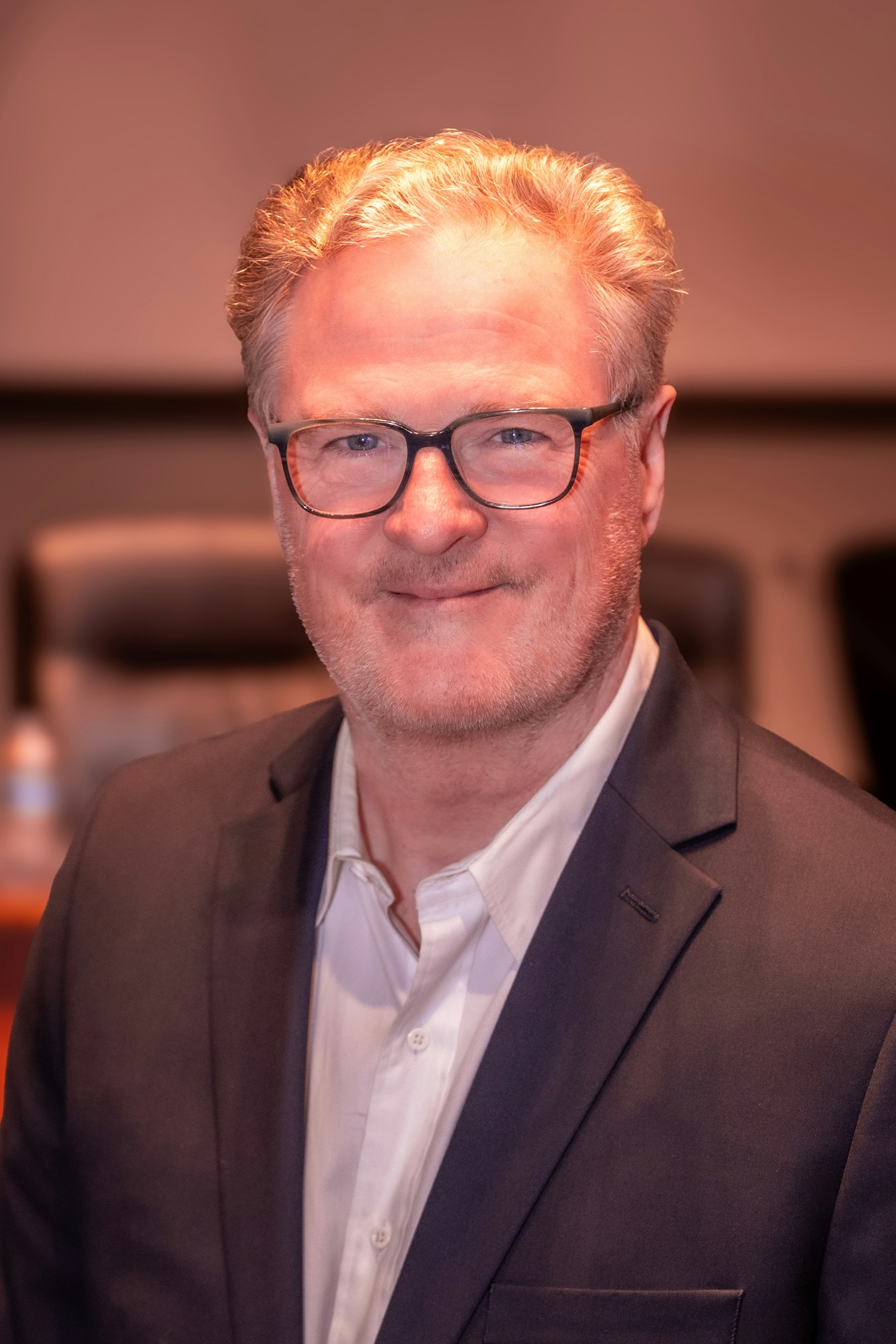 A person wearing glasses and a suit, smiling in a professional setting.