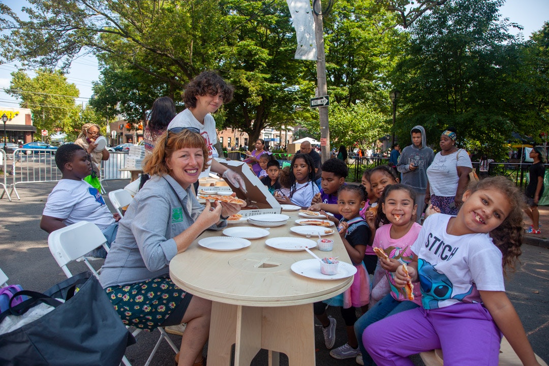 A group of people, including children, enjoying pizza outdoors at a table in a park setting.