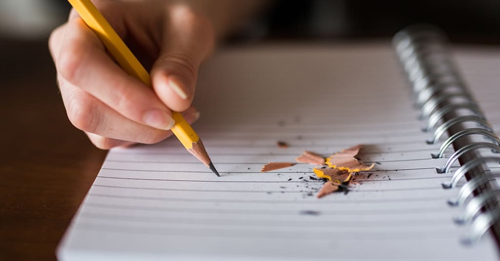 A hand writes with a pencil on lined paper, next to pencil shavings.