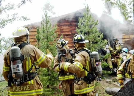 Firefighters wearing protective gear with air tanks preparing to address a house fire, with smoke visible in the background.