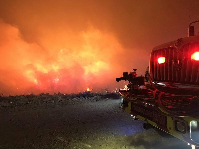 A fire truck faces a large wildfire at night, with intense orange flames and smoke.
