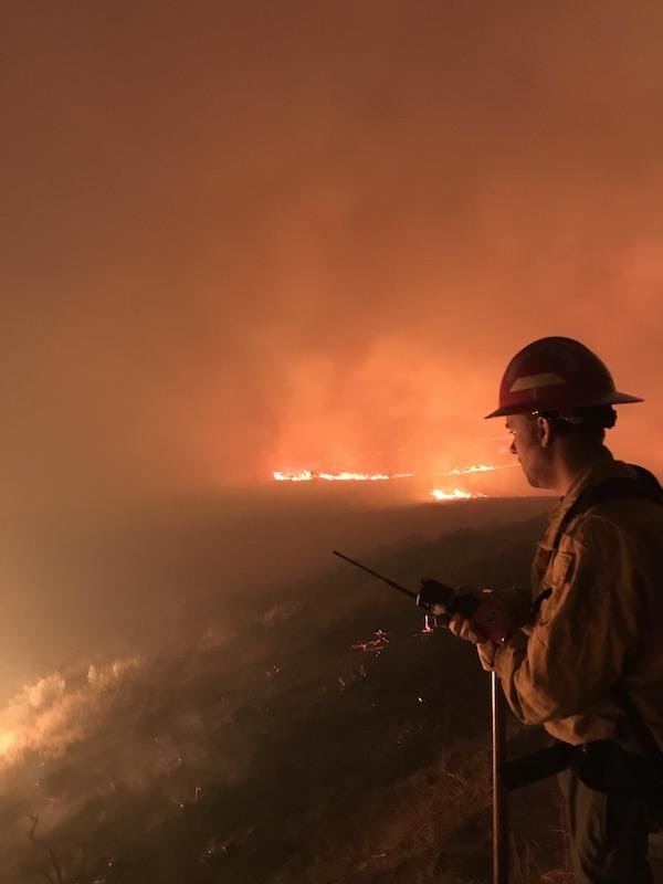 Firefighter watching a wildfire at night, holding a radio, under an orange sky.