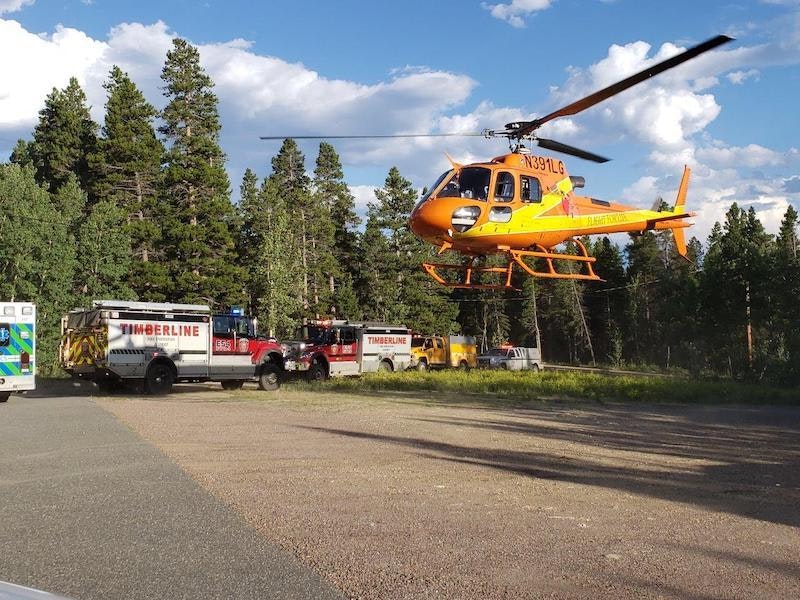 A helicopter hovers near emergency vehicles amidst a forest.