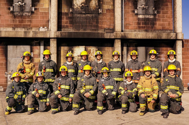 A group of firefighters in uniform posing in front of a charred structure.