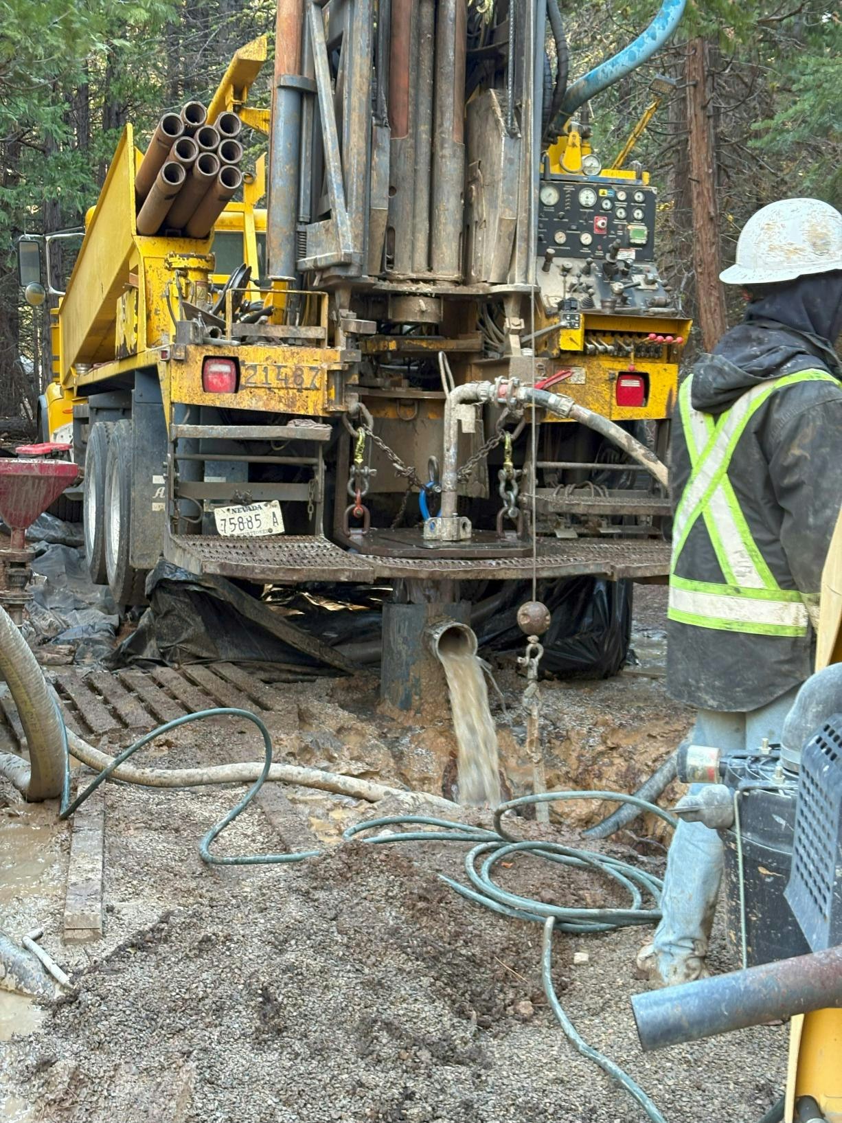 An industrial drilling rig working in a forested area, with pipes, hoses, and a worker observing the operation.