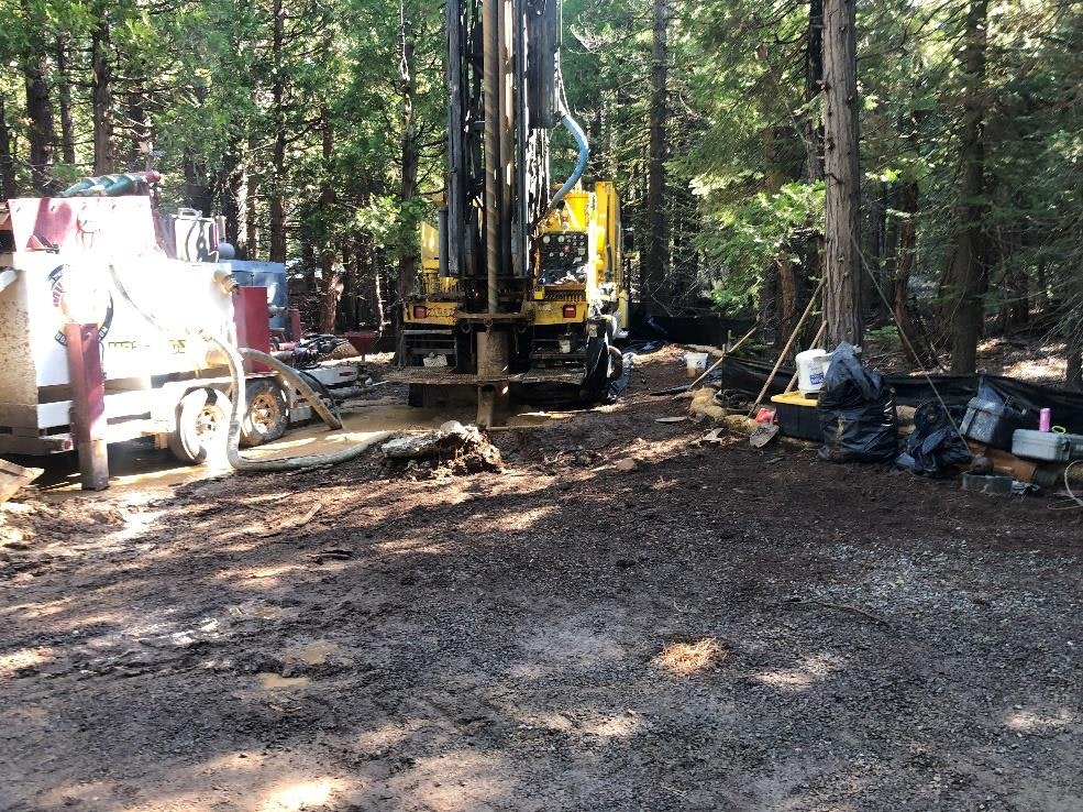 The image shows a drilling rig in a wooded area, surrounded by equipment and materials, likely for a construction or mining project.