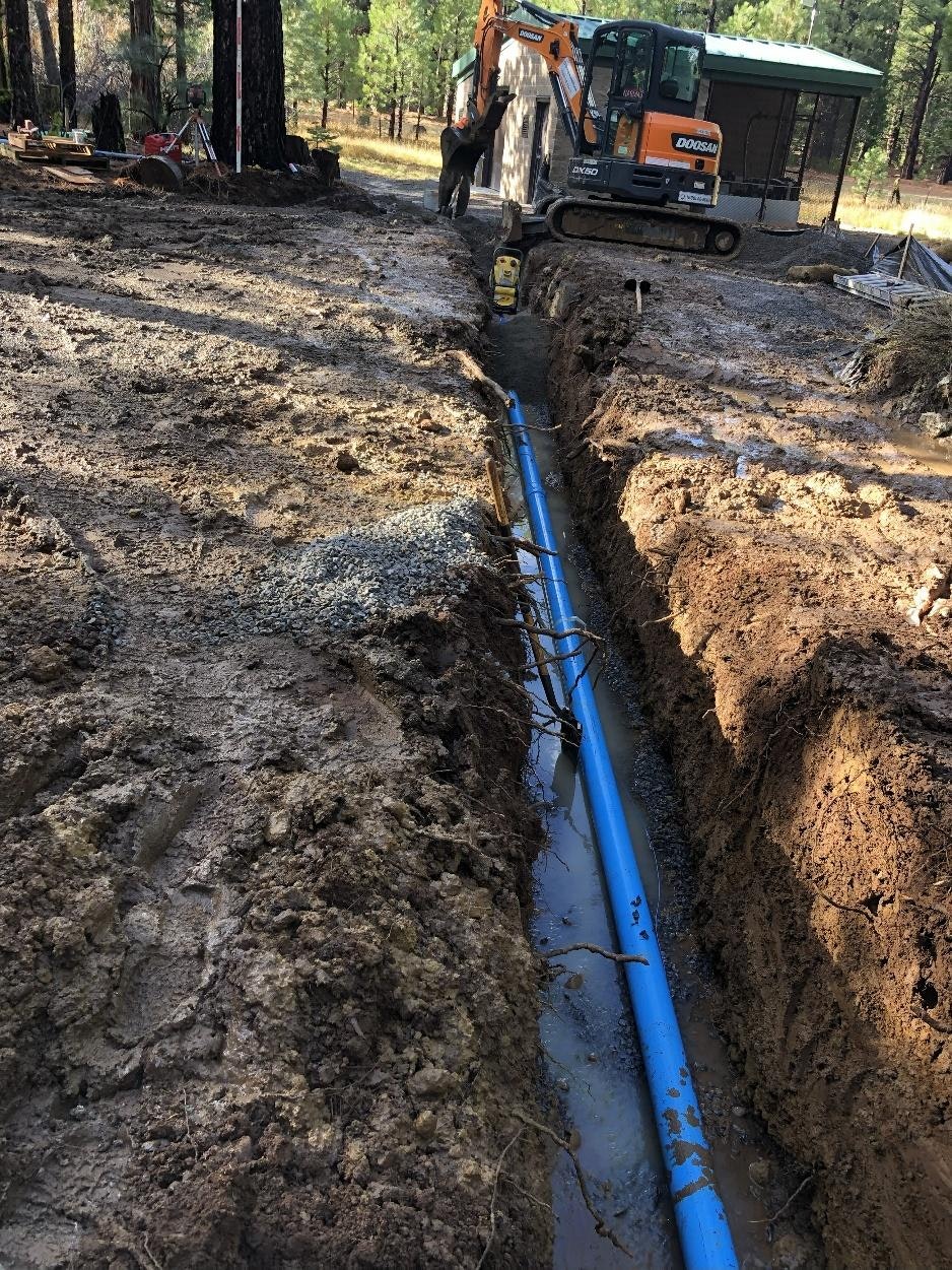 A construction site with a trench dug for plumbing installation, featuring a blue pipe and heavy machinery in a forested area.
