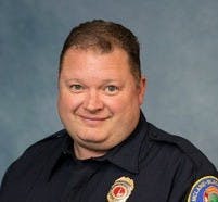 The image shows a person wearing a dark uniform with an emblem, smiling against a blue background.