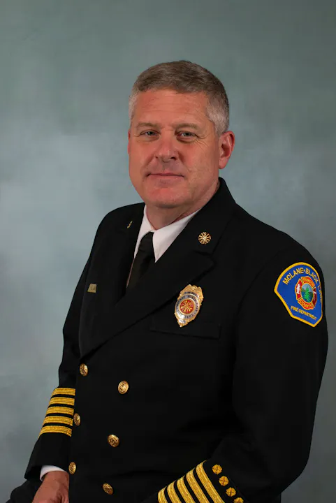 A man in a formal black fire chief uniform with gold accents and badges, posing against a light background.