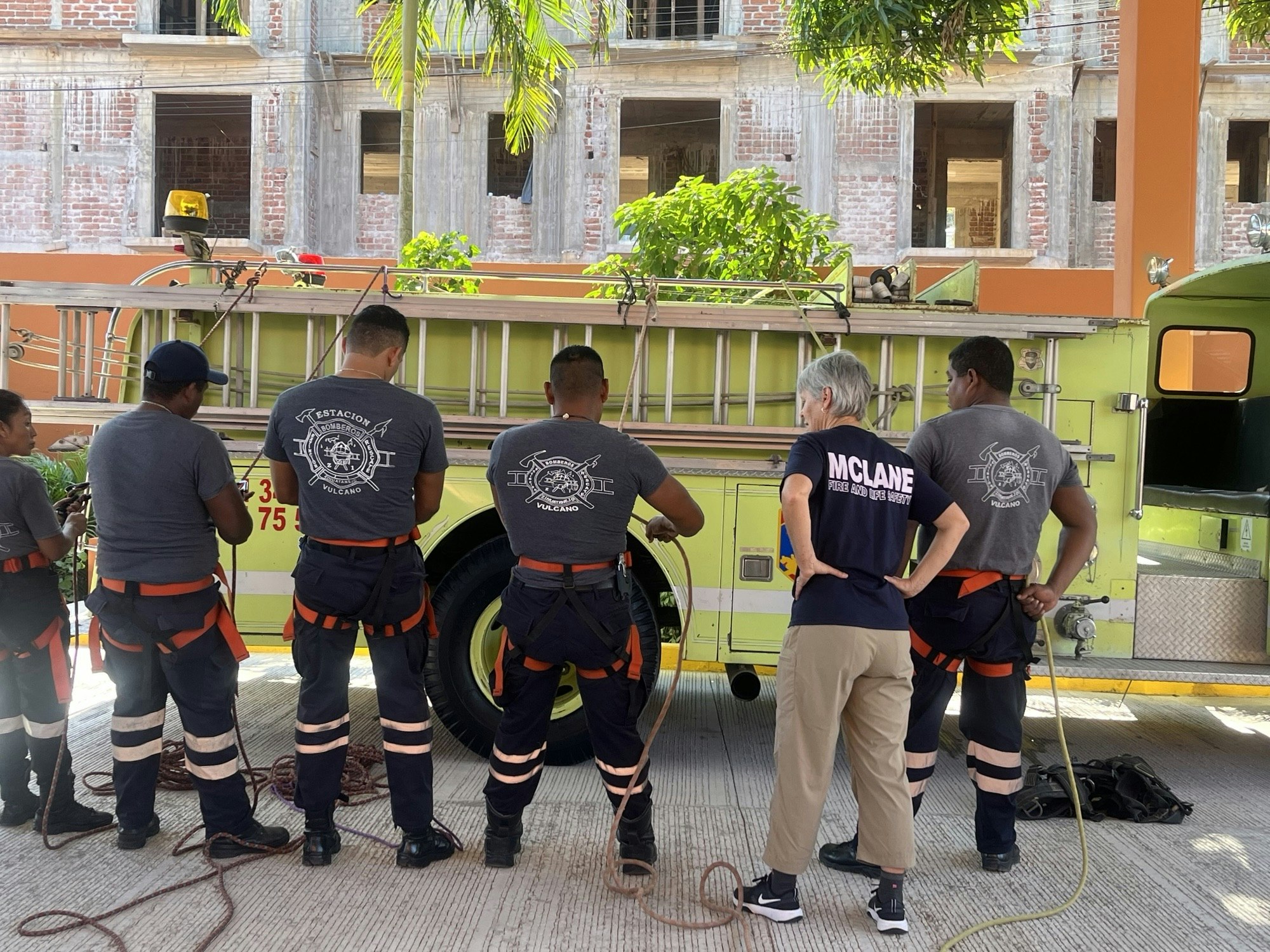 A group of firefighters in uniforms prepares by a fire truck, practicing with ropes, in front of a partially-built structure.